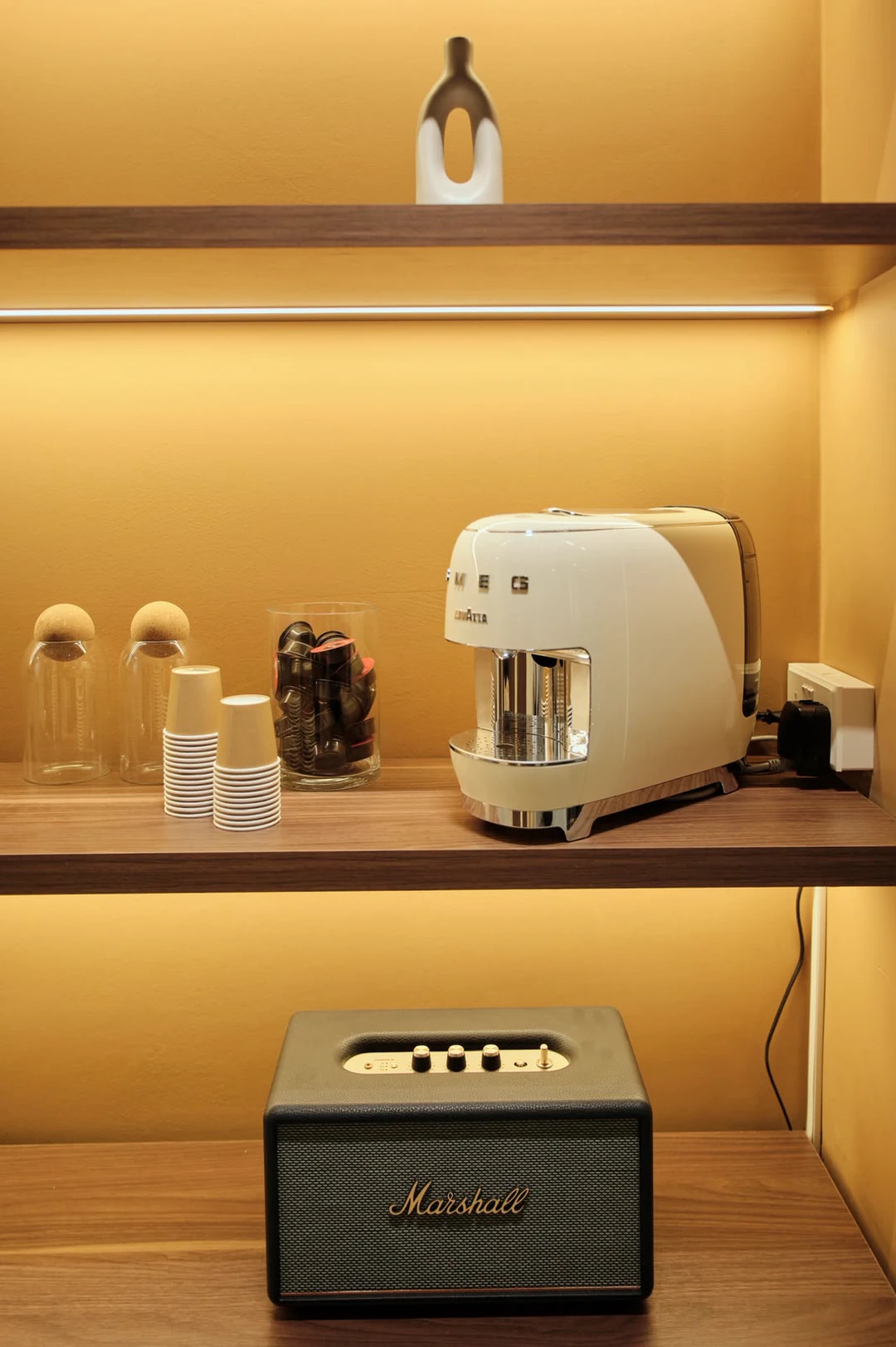 A cream espresso machine and cups on a lit walnut shelf, with a speaker below.